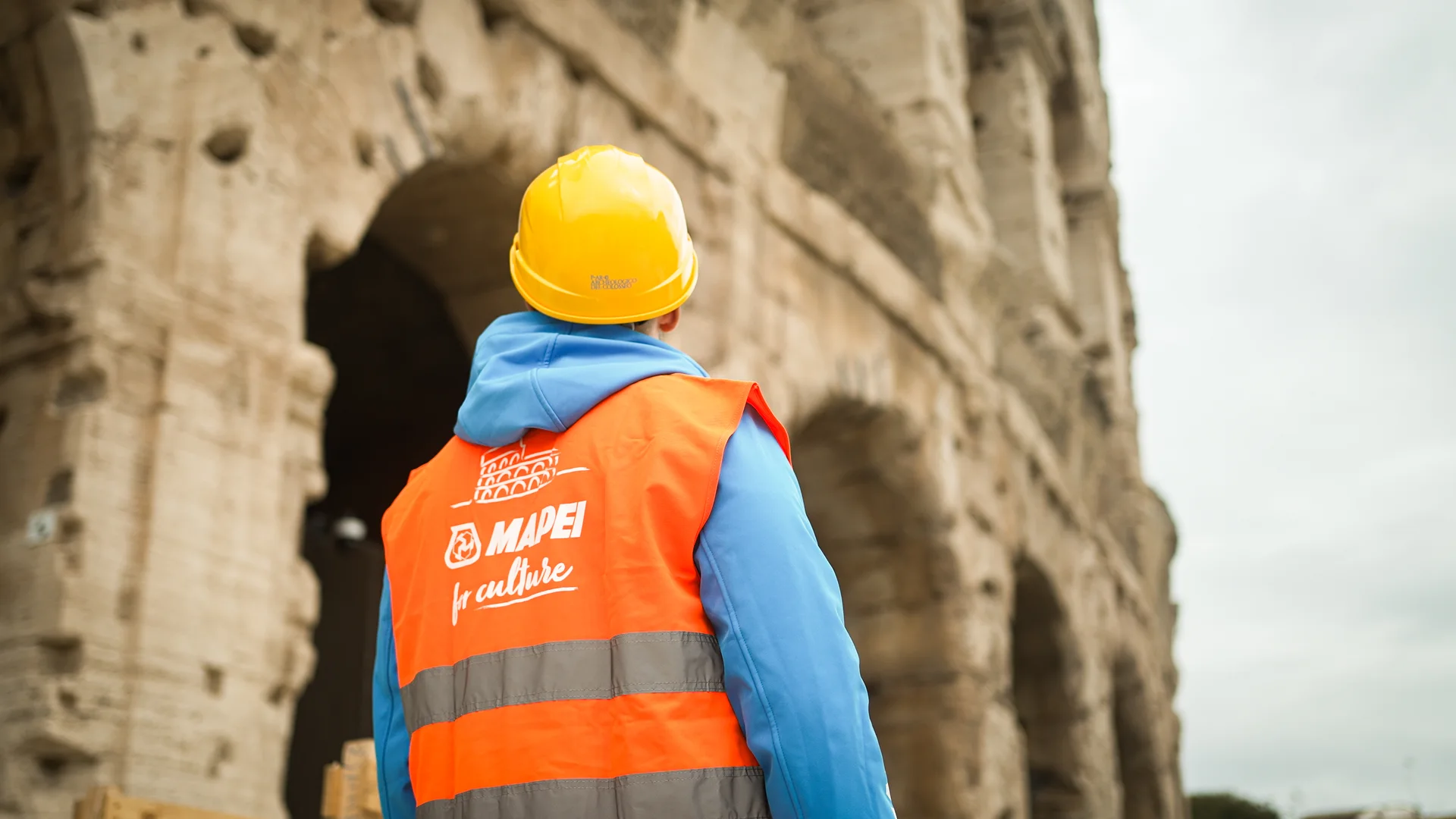 A worker in a Mapei for Culture hi-vis vest facing the ancient arches of the Colosseum